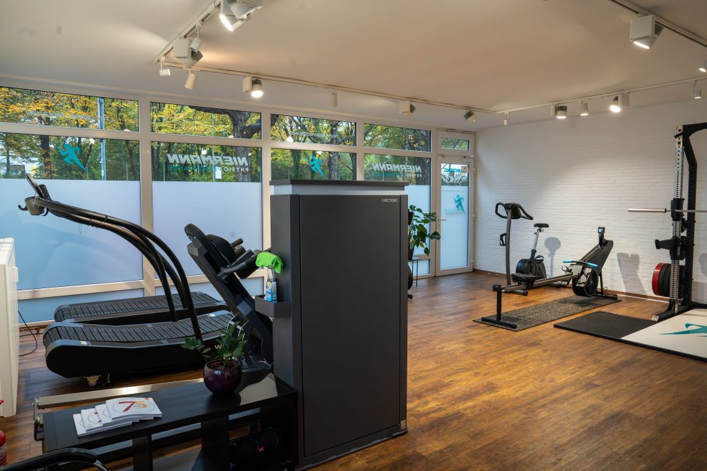 Ein Fitnessraum mit verschiedenen Trainingsgeräten. Im Vordergrund befinden sich zwei Laufbänder und ein Fitnessgerät. Der Raum ist gut beleuchtet, mit Fensteransicht auf die Außenwelt.