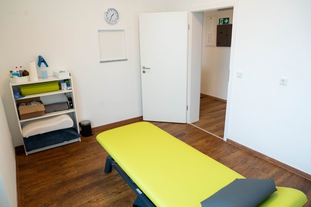 Ein Behandlungsraum mit einer grünen Liege, einem Stuhl und einem kleinen Regal mit verschiedenen Therapiewaren. Der Raum wirkt hell und modern.