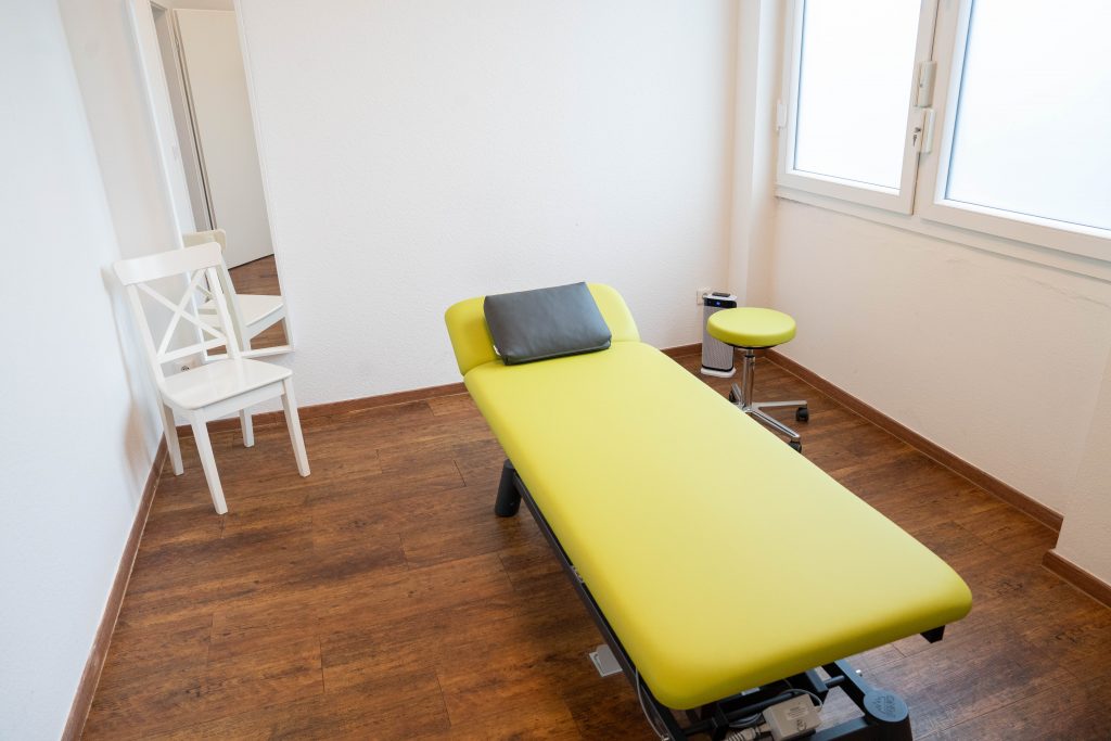 Ein Physiotherapieraum mit einer grünen Liege, einem Stuhl und einem Rollhocker, der Raum ist einfach und modern eingerichtet.