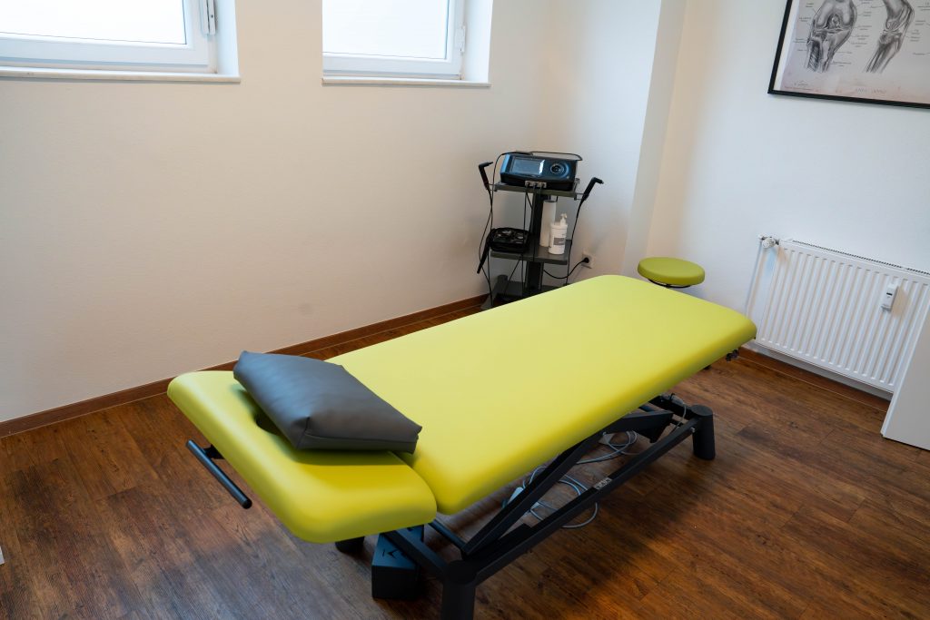 Eine grüne Behandlungsliege in einem modernen Physiotherapieraum mit einem Gerät neben der Liege.