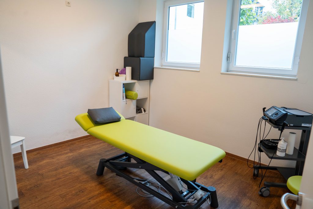 in weiteres Behandlungszimmer mit einer grünen Liege, einem Stuhl und einem Regal mit verschiedenen therapeutischen Materialien.